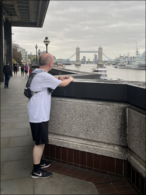 Paul has stopped running - shorts and T shirt and runners backpack with water - looking away from form the camera towards Tower Bridge in the background - cloudy grey sky