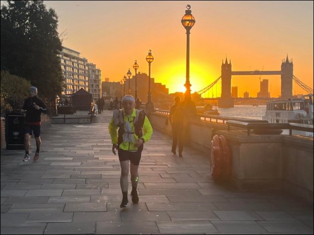 Thames Path next to Old Billingsgate Market, Paul is on the left wearing running kit and a dayglo yellow top, running towards the camera, a magnificent bright sunrise is in the middle, Tower Bridge on the right, the sun is 66% visible and dominates the photo, the temperature is 10°, the cloudless sky is a perfect blue though that is not clear in the photo, a perfect day for a run