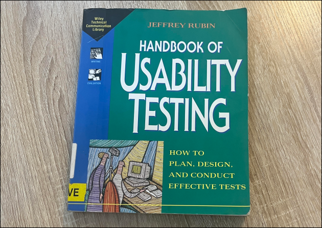 a photo of The Handbook of Usability Testing by Jeffrey Rubin, with a blue and green design on the front cover, sitting on a wood effect desk